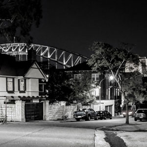View of Kirribilli Avenue at night showing parked cars, and part of the Sydney Harbour Bridge arch above and behind the houses.